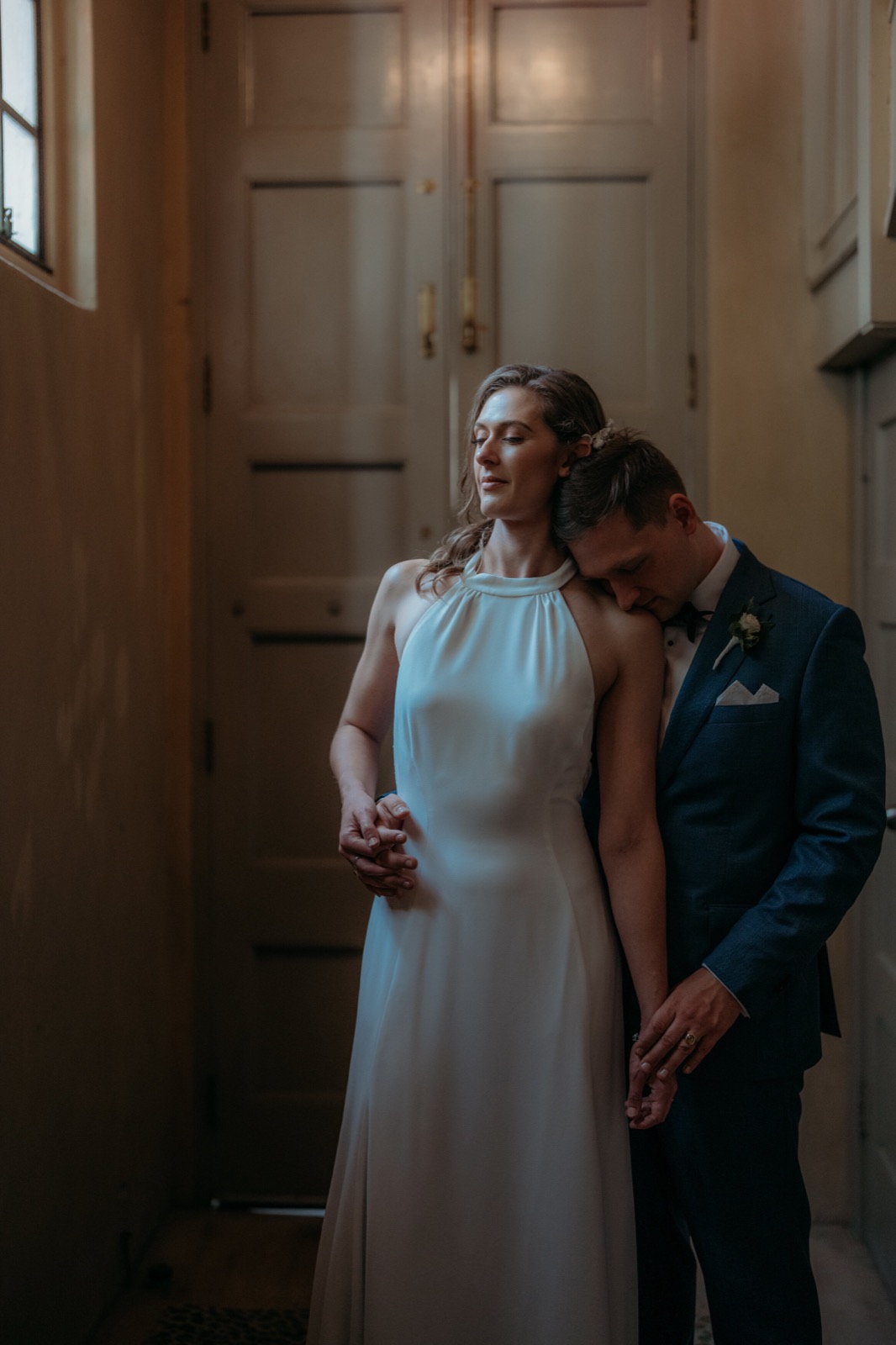 Groom resting his head on his bride's shoulder in a quiet doorway