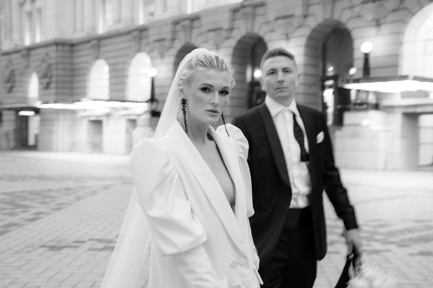 Bride in a white blazer walking with her groom through the city